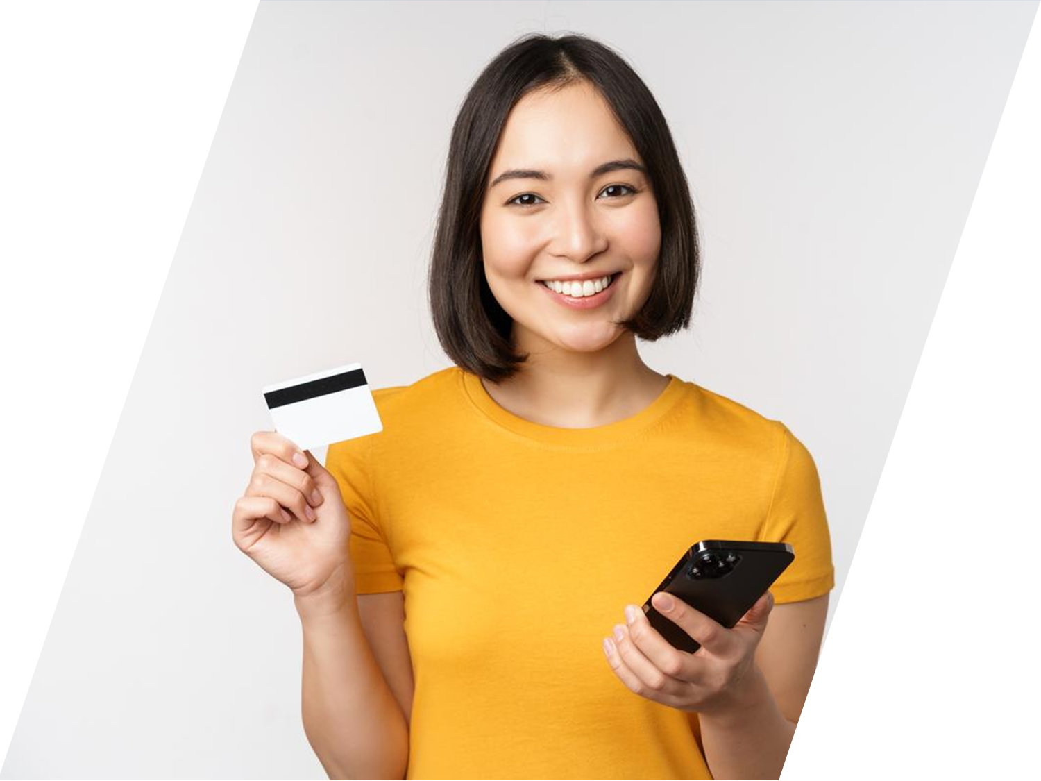 A smiling young woman holds up a credit card in one hand and a smartphone in the other against a white background.