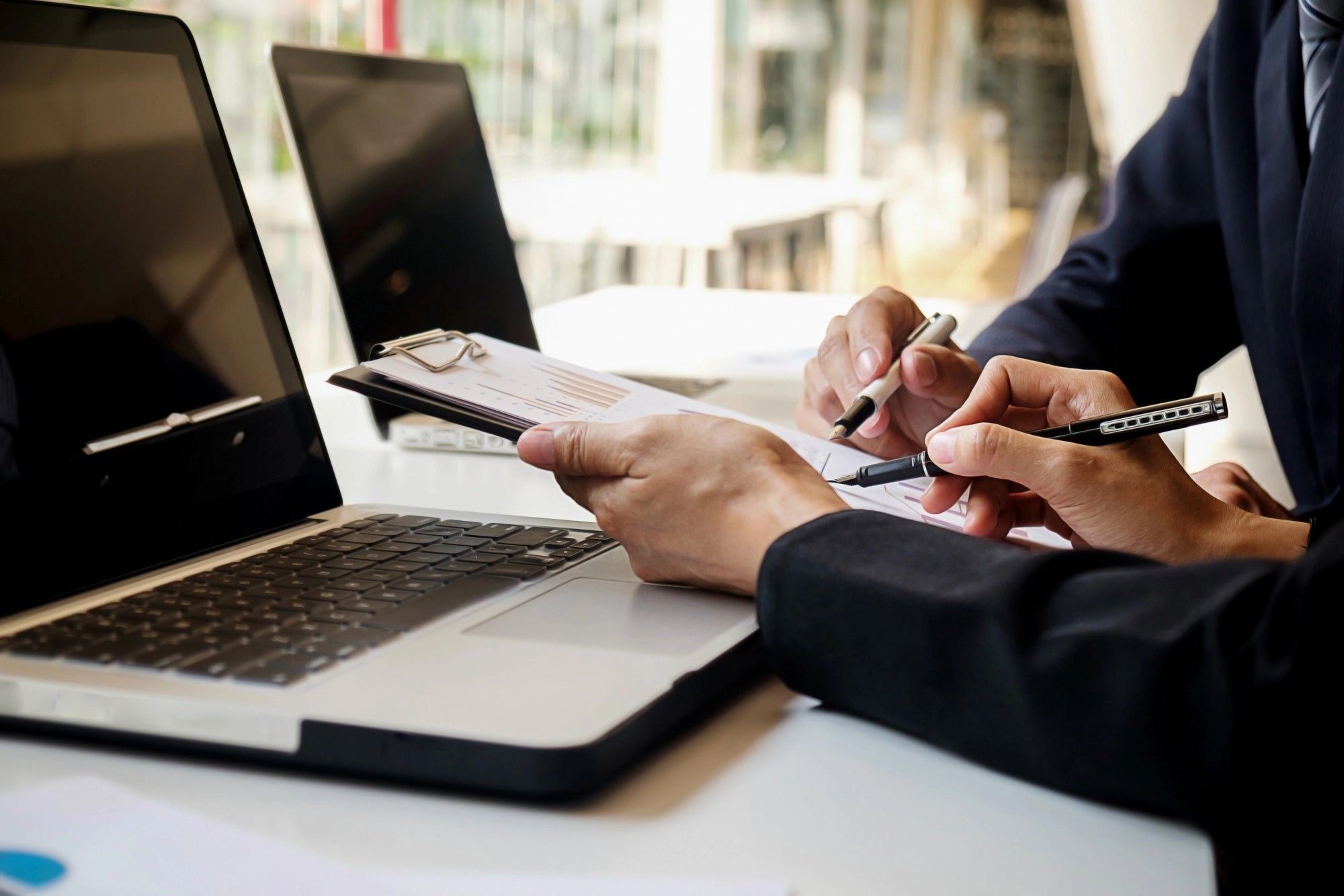 Close-up of two business professionals in suits reviewing data charts on a clipboard with pens next to an open laptop.