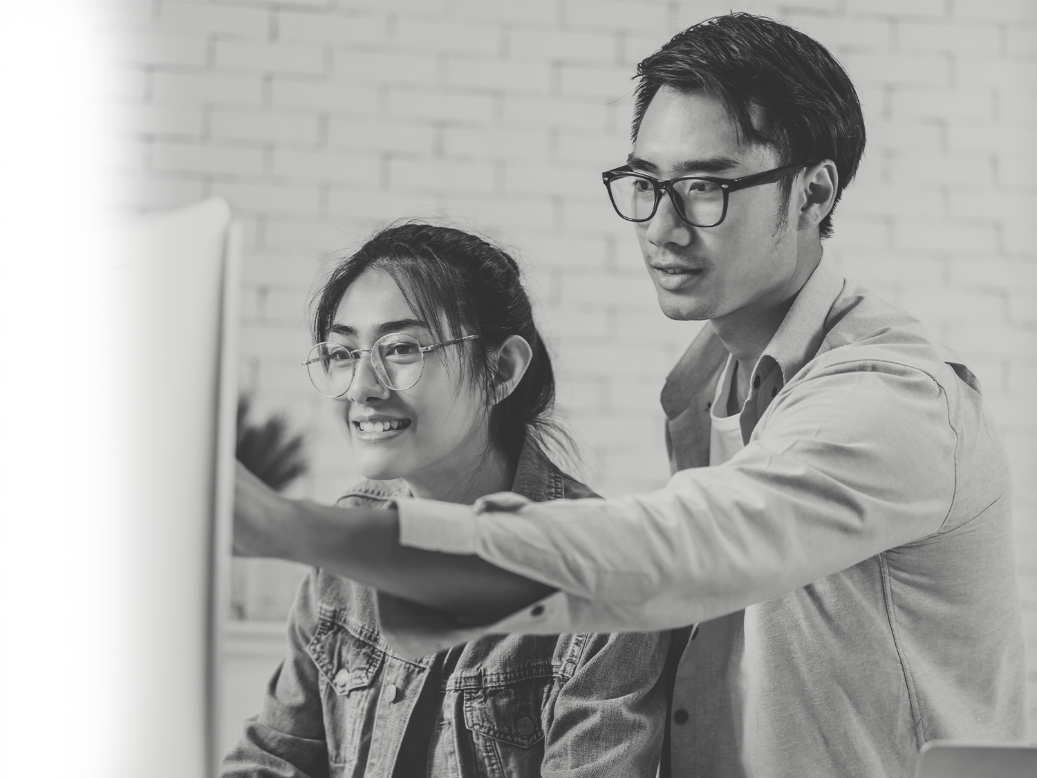 A black and white photo of a smiling young woman with glasses sitting at a desk while a man standing next to her points at a computer screen during a collaborative work session.