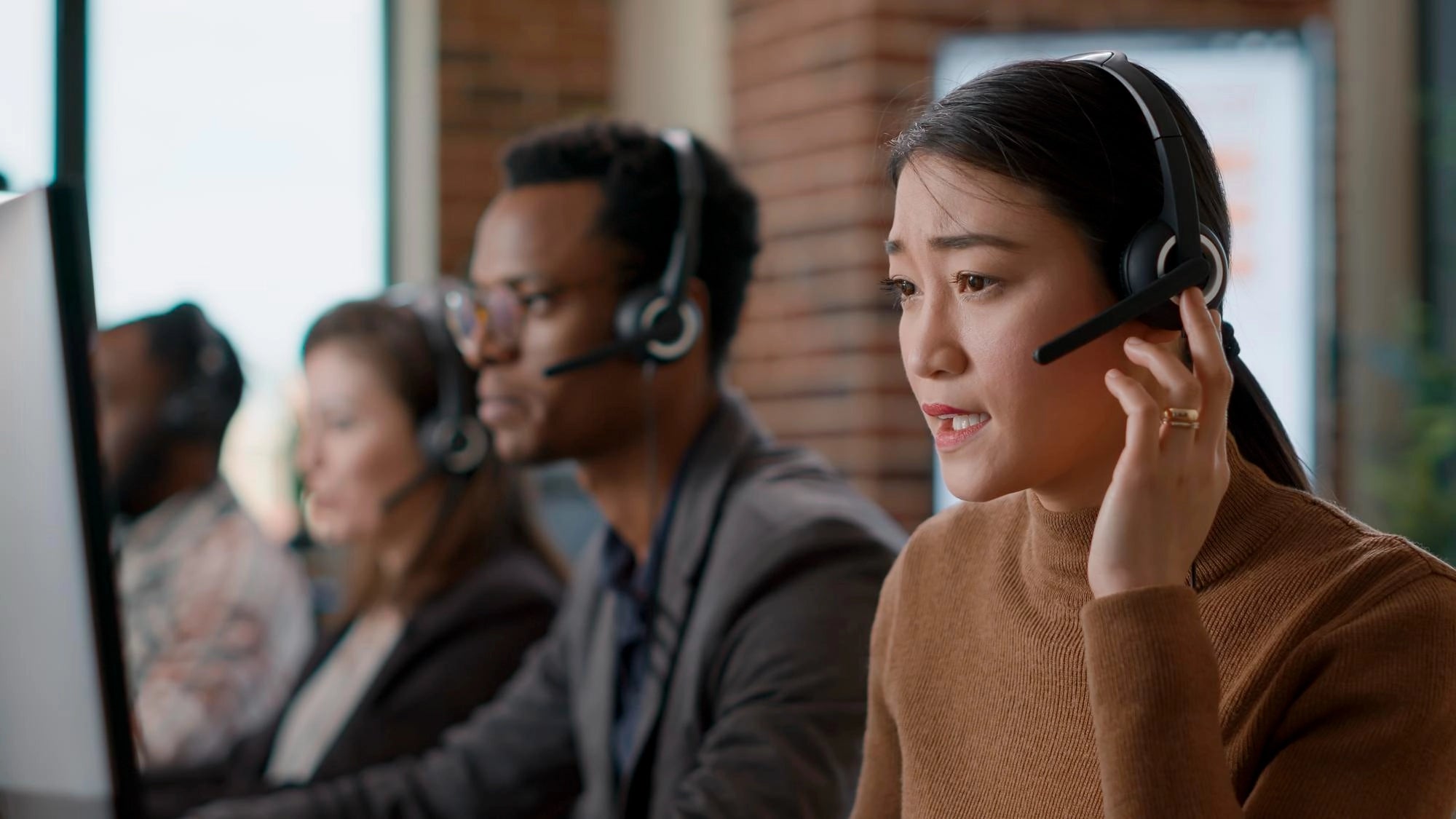 An Asian female customer support agent wearing a headset speaking during a call in a busy office with colleagues in the background.