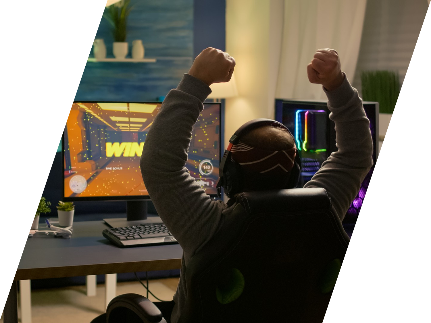 Rear view of a gamer raising his fists in celebration in front of a desktop monitor displaying a video game victory screen.