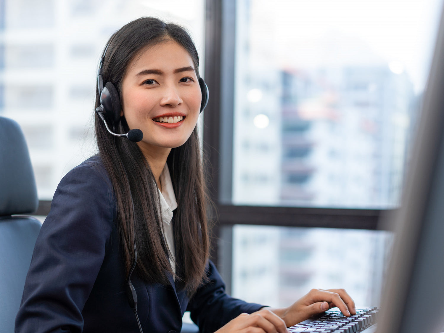 A smiling Asian customer support representative wearing a headset sits at her desk, typing on a keyboard, ready to provide assistance.