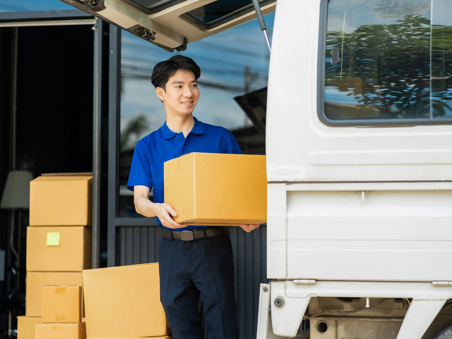 A delivery man in a blue polo shirt and dark pants smiles as he holds a cardboard box, standing next to his delivery van, with a stack of boxes nearby.