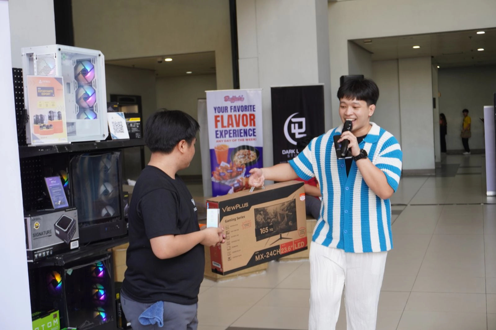 An event host in a blue and white striped shirt presenting a ViewPlus gaming monitor box to a winner at a tech expo.