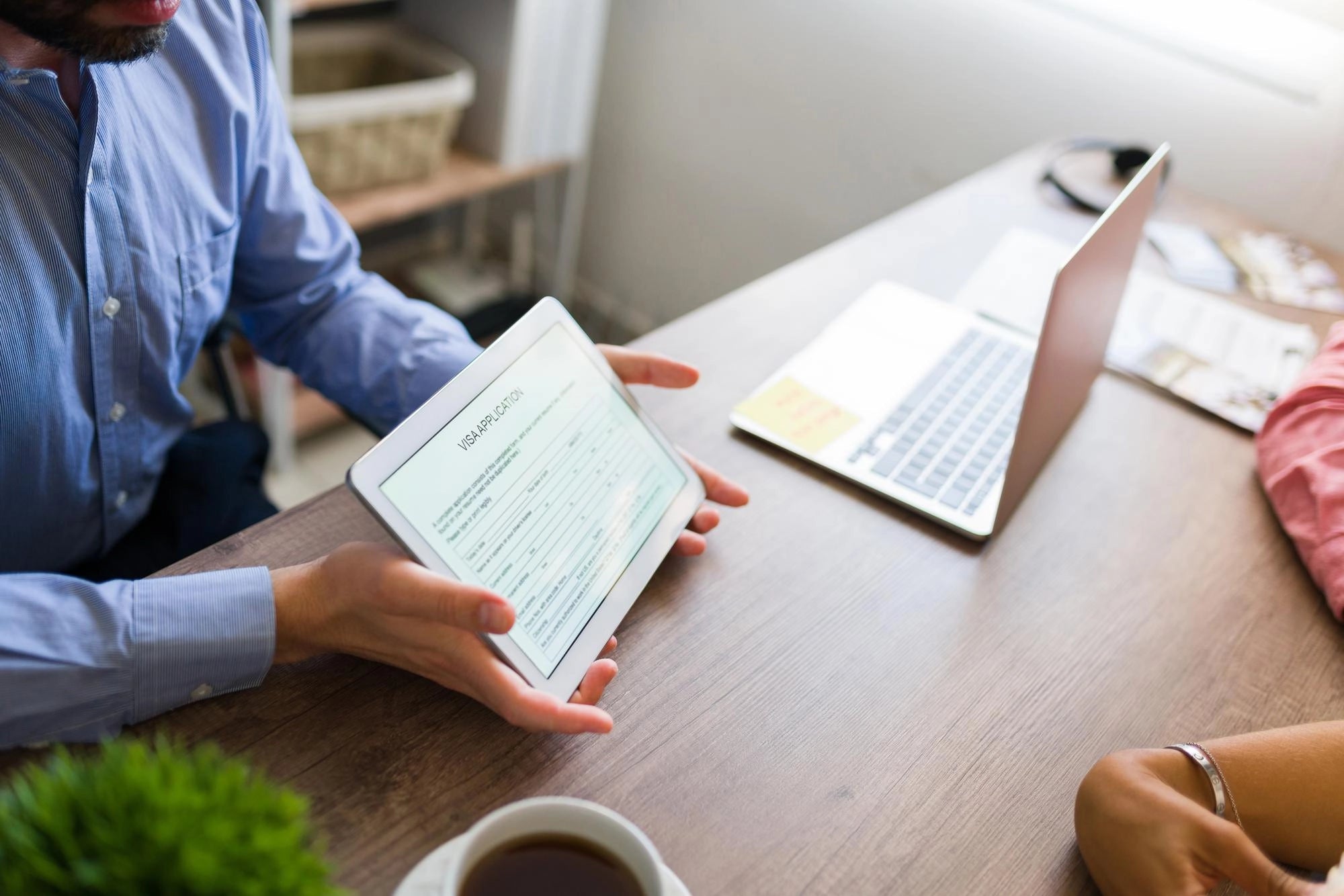 Overhead view of an advisor showing a digital visa application form on a tablet to a client at a wooden desk with a laptop.