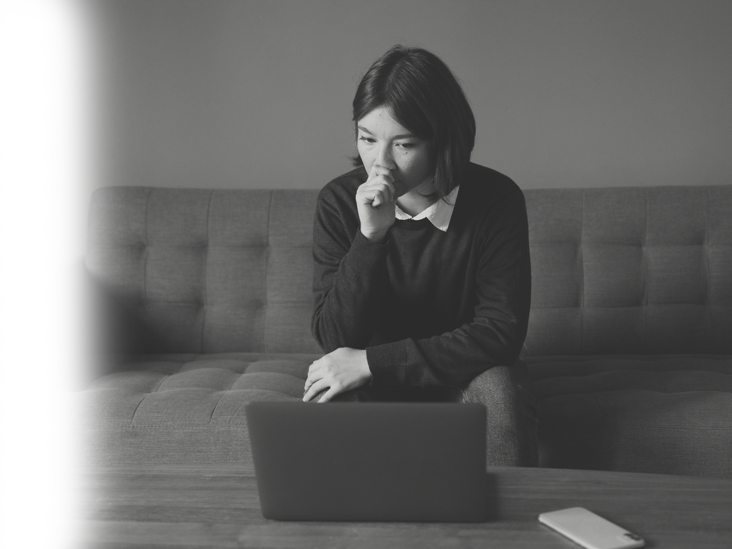 A black and white image of a thoughtful young woman sitting on a sofa and looking intently at her laptop screen placed on a coffee table.